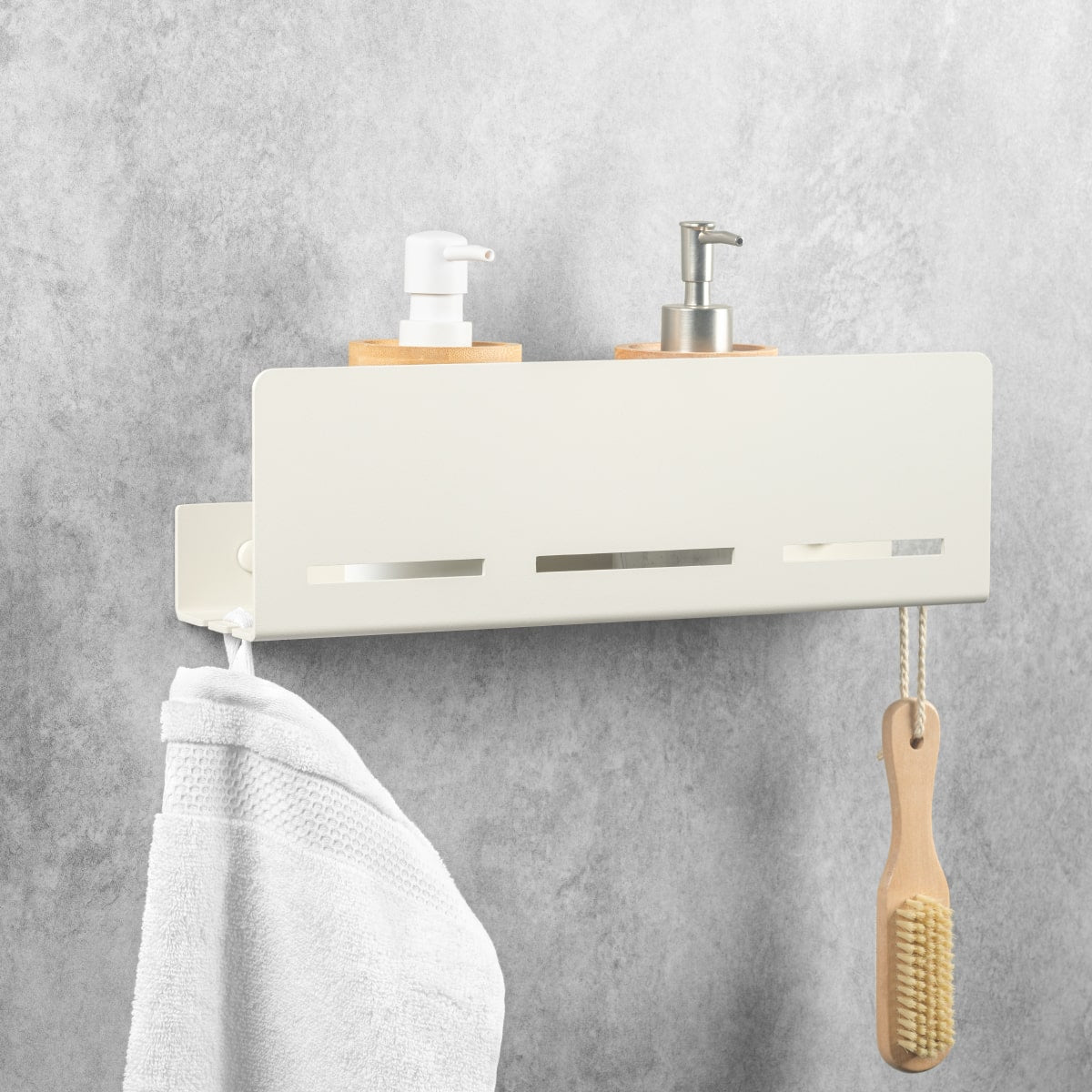 Étagère murale minimaliste en métal beige avec deux distributeurs de liquides, des ouvertures de ventilation et des crochets sur lesquels sont accrochés une serviette et une brosse, installée sur un mur gris