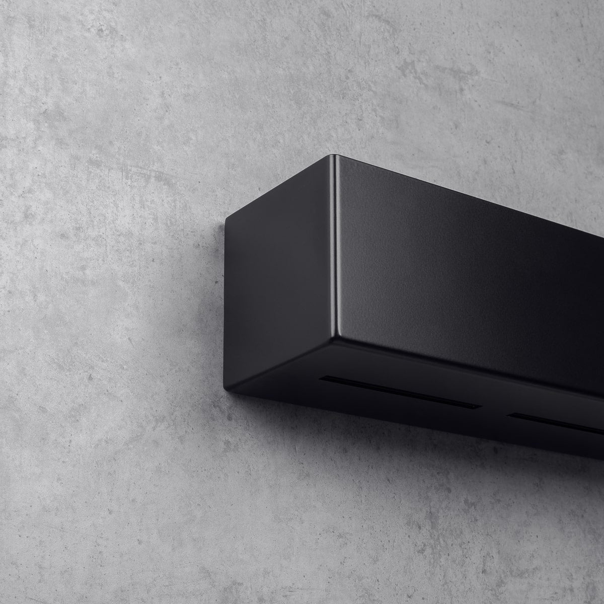 gros plan d'une étagère murale noire au design minimaliste, fixée à un mur gris texturé. Vue de dessous, montrant les fentes de ventilation